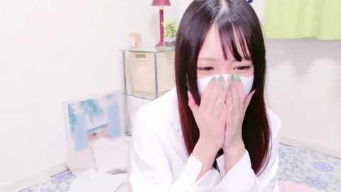 Media: Video of a young Asian woman with long black hair, wearing a white mask, holding her hands in prayer pose, in a white room with a glass shelf and blue-patterned rug.