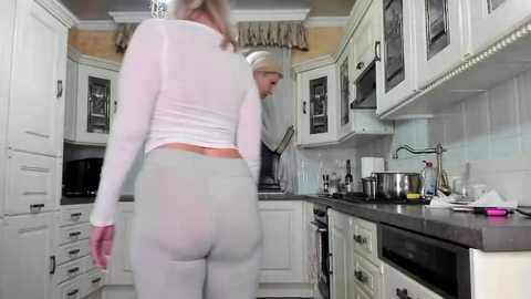 Media: Video of a woman in a white long-sleeve top and grey leggings walking away in a modern kitchen with white cabinets, stainless steel appliances, and a sink.