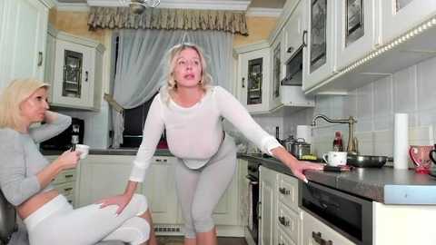 Media: Video of two women in white pajamas, one leaning on a kitchen counter, the other seated. Background features white cabinets, a sink, and a window with blue curtains.