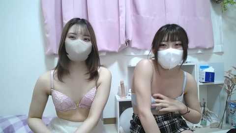 Media: Video of two young East Asian women in matching pink face masks and pastel lingerie, sitting in a bedroom with pink curtains and a white wall.