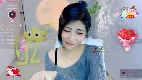 Media: Video of a smiling Asian woman with dark hair, wearing a gray sweater, in a light-colored room with a yellow toy bear and heart-shaped decorations.