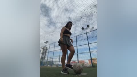 Media: Video of a young woman with medium build and long dark hair in a black sports bra and grey skirt, kicking a soccer ball on a green field, with a blue fence and cloudy sky in the background.