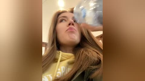 Media: Video of a Caucasian woman with long brown hair, wearing a yellow hoodie, being sprayed with water from a plastic bottle, indoors, blurred background.