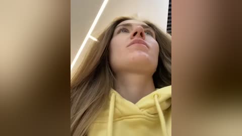 Media: Video of a young Caucasian woman with long, straight brown hair, wearing a yellow hoodie, standing indoors with a neutral background.