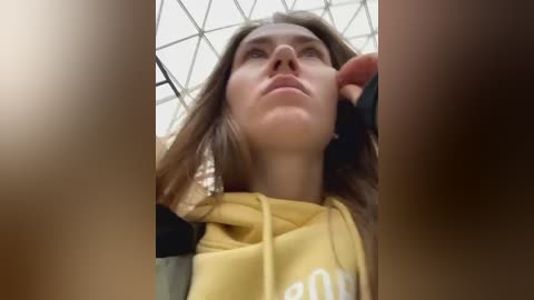 Media: Video of a young woman with long brown hair, wearing a yellow hoodie, gazing upwards, possibly in an urban setting with a grid-patterned ceiling.