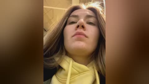 Media: A close-up video of a young woman with straight, shoulder-length brown hair, fair skin, and light makeup, wearing a yellow hoodie, taken in an indoor setting.