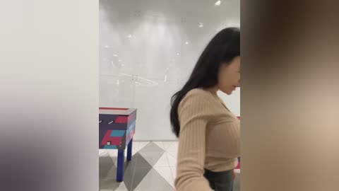 Media: Video of an Asian woman with long black hair in a beige ribbed sweater, walking away from a colorful, geometrically patterned table with chairs in a modern, minimalist room with white walls and a tiled floor.