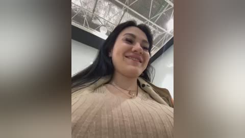 Media: Video of a smiling Latina woman with long black hair, wearing a beige sweater and necklace, standing in an indoor setting with a metallic ceiling.