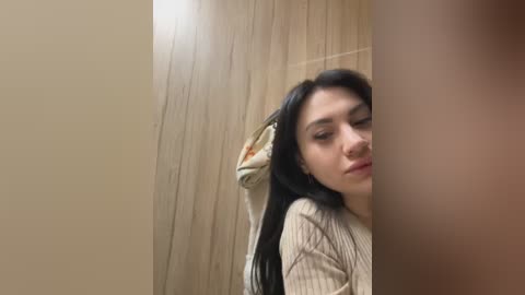 Media: Video of a young woman with long black hair, light skin, and a fair complexion, wearing a beige sweater, standing in a wooden-paneled bathroom, with a blurred man's leg in the foreground.