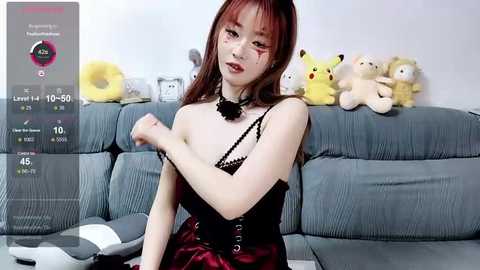 Media: Video of an East Asian woman with fair skin, wearing a black corset and red lipstick, posing on a gray sofa with plush toys.