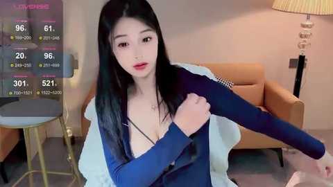 Media: Video of an East Asian woman with long black hair, wearing a blue top, adjusting her sleeve in a modern living room.