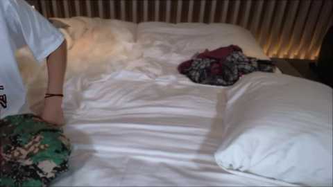 Media: A video of a bed with white sheets and pillows, featuring a partially visible person in a white shirt and camouflage shorts, sitting on the bed. A floral-patterned garment lies on the pillow.