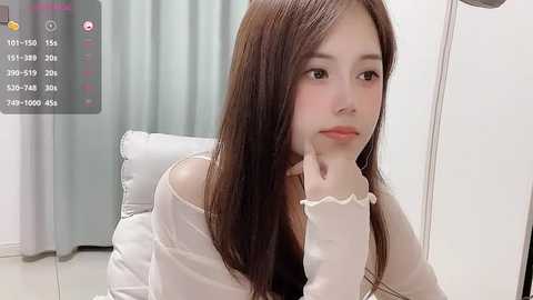 Media: Video of an East Asian woman with long, straight brown hair, wearing a white off-shoulder top, sitting pensively on a white couch. Background features light curtains and a gray wall.