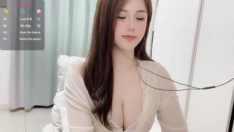 Media: Video of an Asian woman with long brown hair, fair skin, and a slender physique, wearing a sheer, white, lace-trimmed robe, sitting on a bed, eyes closed, headphones on, in a white room.