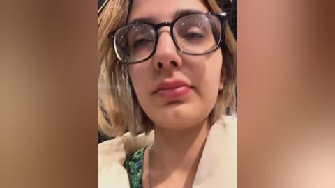 Media: Video of a young woman with light skin and short blonde hair, wearing black-rimmed glasses, a green blouse, and a white sweater, with a neutral expression.