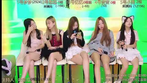 Media: Video of five young Asian women in casual outfits, seated on white chairs against a colorful backdrop, holding mobile phones.