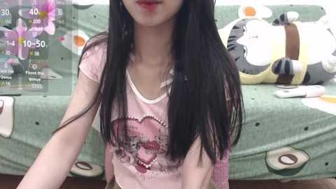 Media: Video of a young East Asian woman with long black hair, wearing a pink T-shirt with a cartoon skull design. Background includes a green couch with plush toys and a digital clock.