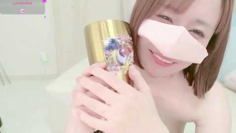 Media: Video of a smiling Asian woman with light skin and shoulder-length brown hair, holding a gold trumpet with a pink bandage over her nose. She's topless, indoors, with a white background.