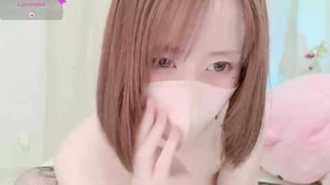 Media: A close-up video of a light-skinned, young woman with straight, shoulder-length brown hair, wearing a pink face mask, partially covering her mouth. The background features a soft, pastel-colored room with minimal details.