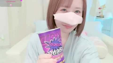 Media: A video of a young Asian woman with straight, light brown hair, wearing a light blue robe, holding a purple \"100%\" face mask box, in a brightly lit, minimalistic room with soft pastel colors.