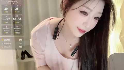 Media: A video of an East Asian woman with long dark hair, light skin, and wearing a light pink top. She's indoors, leaning forward with headphones on, smiling softly.