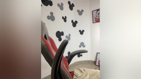 Media: Video of a minimalist room with a red and grey ergonomic office chair, white walls adorned with black and grey Mickey Mouse stickers, and a framed cartoon print on the right.