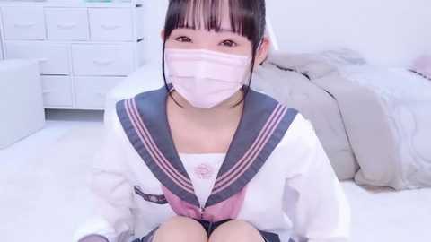 Media: Video of an East Asian woman with a light complexion, wearing a pink surgical mask, a white sailor school uniform, and sitting on a white bed, with white drawers and a grey blanket in the background.