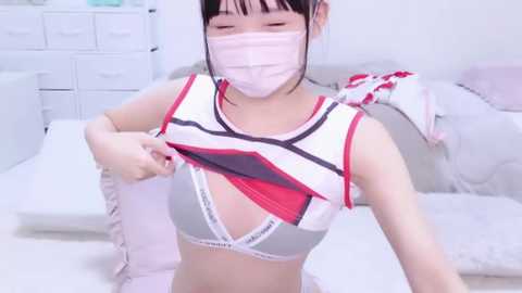 Media: A video of an East Asian woman in a white and red striped crop top, revealing her black bra and midriff, wearing a pink face mask, seated on a white bed with white dresser in the background.