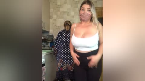 Media: Video of a plus-size woman in a white tank top and black leggings standing in a kitchen, wearing a pink face mask, while a woman in a polka-dot apron cooks in the background.