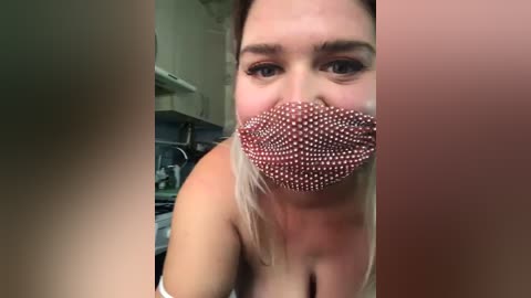 Media: Video of a blonde woman with fair skin, wearing a red, polka-dot face mask, in a kitchen setting, featuring a blurred background.