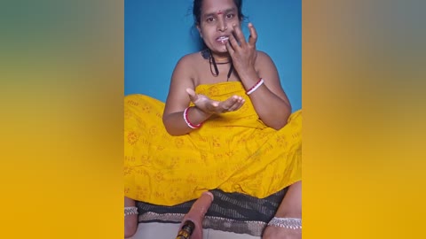 Media: Video of a South Asian woman with medium skin tone, wearing a bright yellow sari, sitting on the floor with a blue background. She is holding her finger to her mouth in a playful manner.