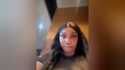 Media: Video of a young Black woman with long black hair, wearing a green shirt, looking into a mirror with a blurred background of a wooden ceiling.