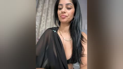 Media: A video of a young South Asian woman with long black hair, wearing a sheer black shawl, revealing a black bra, and a gold chain necklace. She stands against a blurred background with a patterned curtain.