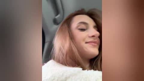 Media: Video of a young woman with shoulder-length, light brown hair, wearing a white, fluffy sweater, smiling gently with closed eyes, sitting in a car.