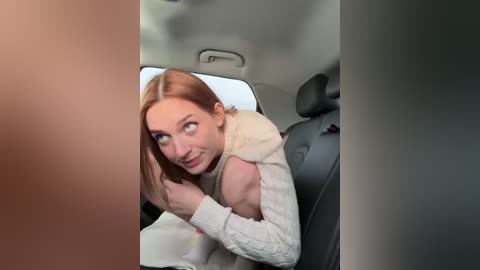 Media: Video of a young woman with fair skin and straight, shoulder-length auburn hair, wearing a cream-colored sweater, sitting in a car, looking directly at the camera.