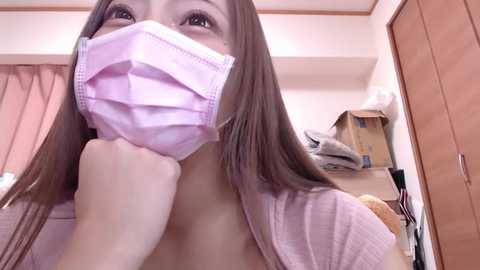 Media: Video of a young Asian woman with straight brown hair, wearing a light pink mask and a light pink cardigan, looking contemplative. Background shows a cluttered room with wooden furniture, boxes, and a teddy bear.