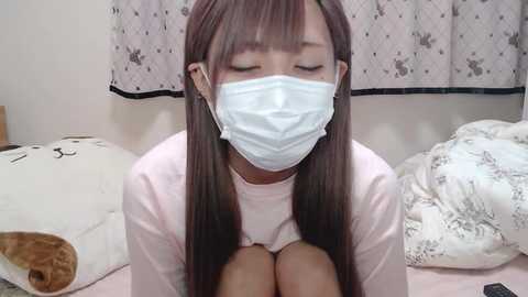 Media: A video of an Asian woman with long brown hair, wearing a white mask, pink top, and patterned bedding in a simple bedroom.