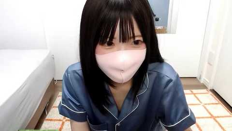 Media: Video of an Asian woman with long black hair, wearing a blue silk pajama shirt and pink surgical mask, kneeling on a patterned rug in a brightly lit bedroom with white walls and a bed.