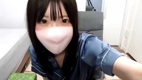 Media: A video of an Asian woman with long black hair, wearing a blue denim shirt and a white surgical mask, taking a selfie in a bright, modern bedroom with wooden floors.