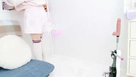 Media: Video of a young girl in a pastel pink outfit and white knee-high socks, standing in a minimalist, white room with a pink vibrator and a blue cushion.