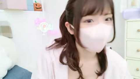 Media: A video of a young Asian woman with fair skin, wearing a white mask, pastel-colored hair, and a pink robe, standing in a pastel-colored room with Halloween decor.