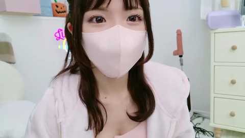 Media: Video of an Asian woman with long brown hair, wearing a pink mask, pink hoodie, and pink top, in a pastel-colored bedroom with a toy pumpkin and a white dresser.