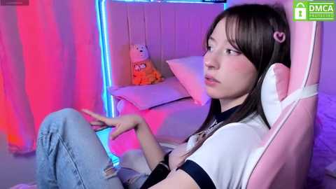 Media: Video of a young Asian woman in a pink gaming chair, wearing a white t-shirt and blue jeans, with a cute plush toy and glowing neon lights in the background.