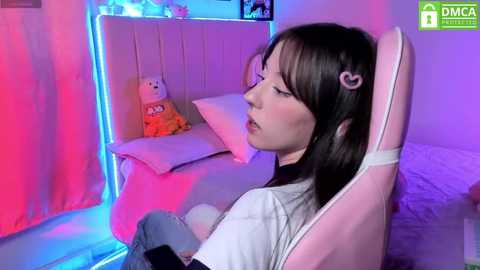 Media: A video of a young Asian woman with straight black hair, sitting in a pink gaming chair, wearing a white t-shirt, in a dimly lit, colorful bedroom with plush toys and neon lights.