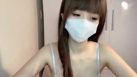 Media: Video of an East Asian woman with long brown hair, wearing a white surgical mask, a white spaghetti-strap tank top, and a light pink background.