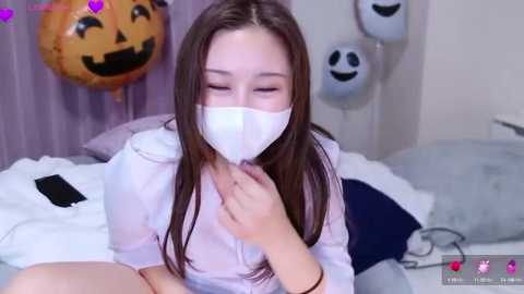 Media: A video of an Asian woman with long brown hair, wearing a white mask, lying on a bed with a Halloween-themed balloon and white pillow.