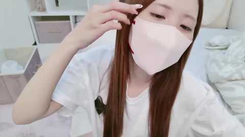 Media: Video of an East Asian woman with long straight brown hair, wearing a white mask, adjusting her hair in a bedroom with light-colored furniture and a bed.