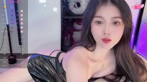 Media: Video of an East Asian woman with long black hair and fair skin, wearing a shiny black dress, seated indoors with a panda plush toy in the background.