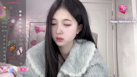 Media: Video of a young Asian woman with long black hair, wearing a fluffy gray robe, indoors. Background includes a door, pink balloons, and \"Happy Valentine's Day\" text.