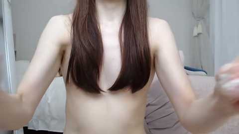 Media: Video of a nude, fair-skinned, slender young woman with long, straight brown hair parted down the middle, standing indoors with white walls and a bed in the background.